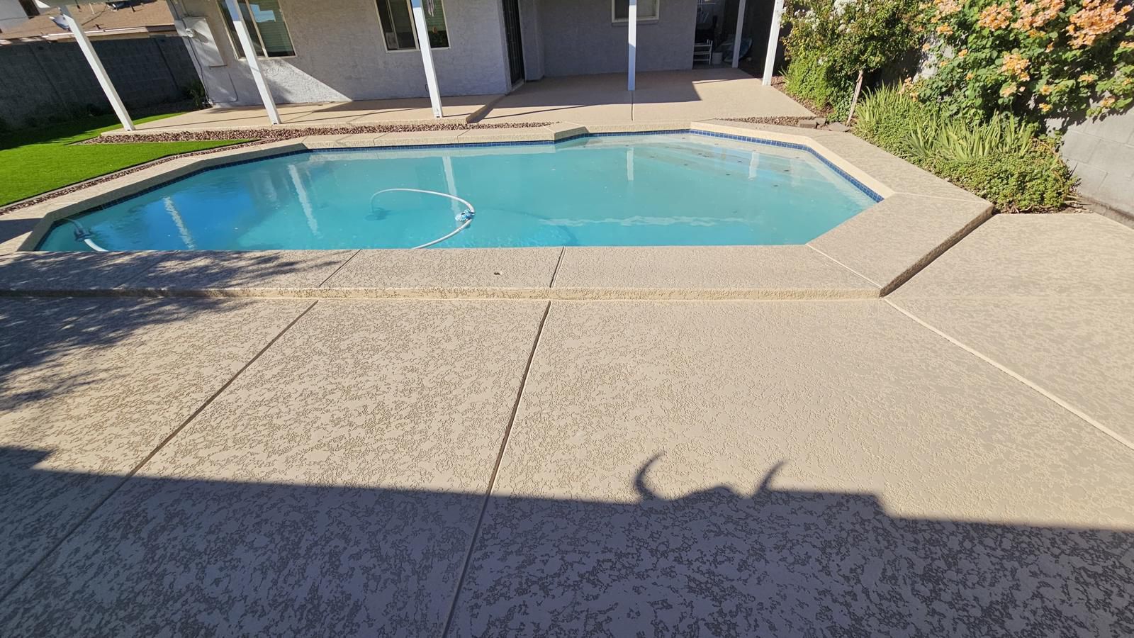 Concrete pool deck next to a swimming pool and a manicured lawn. The deck has a speckled texture.