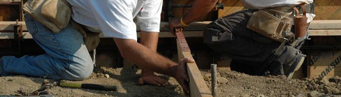 Two construction workers kneeling, working with wood. One holds a level. Outdoors.