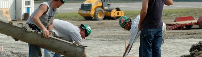 Construction workers pouring concrete on a job site. Yellow tractor visible in the background.