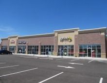 A strip mall with businesses, brick exterior, and a large parking lot on a clear day.