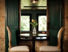 Two chairs face a small table with flowers, framed by a window and green shutters against a brick wall.
