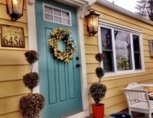 Teal front door with wreath and topiary flanking a yellow cottage; brass lanterns.