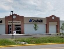 Exterior of a Goodwill store; tan and brick facade, blue Goodwill logo, grass and parking area.
