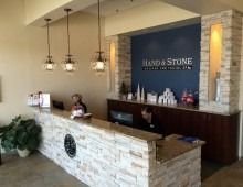 Reception area of a Hand & Stone spa. A staff member is at the reception desk, and there are pendant lights above.