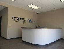 Reception area with a white desk, IT XCEL Consulting logo, and a ceiling light.