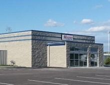 A light brick commercial building with glass entrance, dark trim, and a sign under a blue sky.