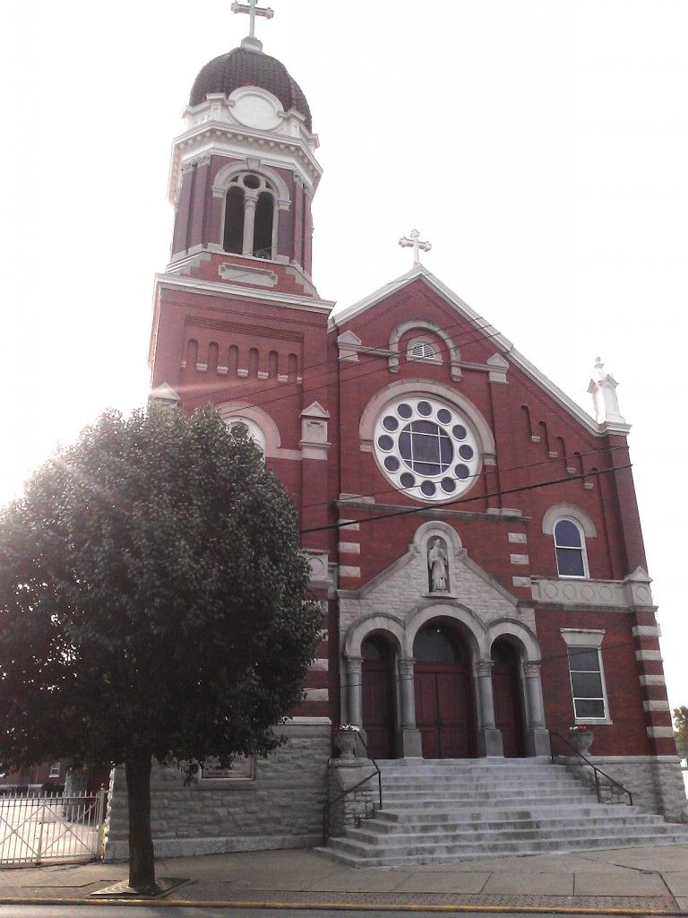 Red brick church with a tall tower, steps, and a large tree in front.