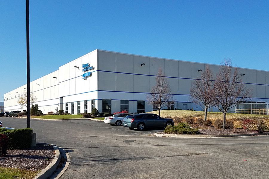 Large white warehouse building with blue logo, parked cars, clear blue sky.