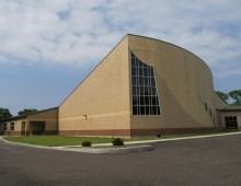 Beige brick building with a curved wall and a large, angled window. Asphalt in front, blue sky.