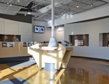 Modern showroom with wood flooring, light-colored walls and displays. A central, interactive display is angled in the center of the room.