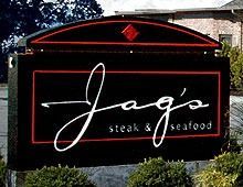 Sign for Jag's Steak & Seafood: black background, red trim, white cursive text, red diamond, outdoors.