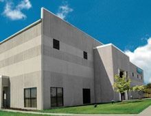 Gray industrial building with small windows against a blue sky, green grass in front.