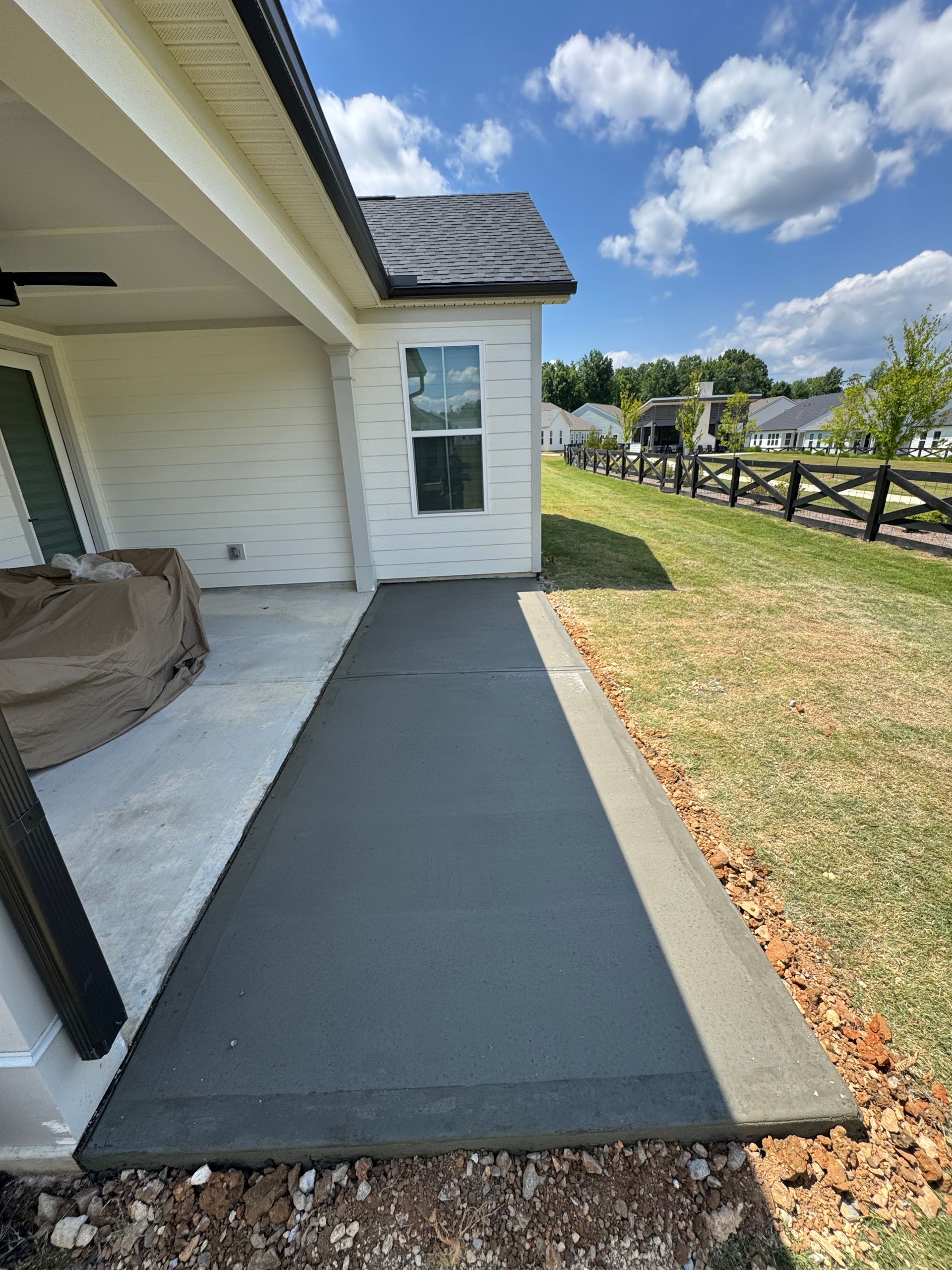 Freshly poured concrete walkway beside a white house, leading past a grassy yard and wooden fence.