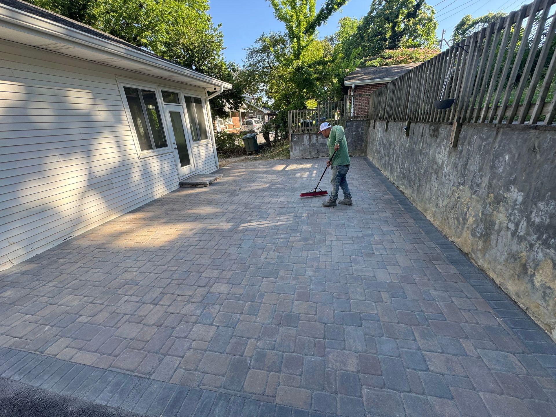 Person sweeping a brick-paved driveway beside a house and stone wall.