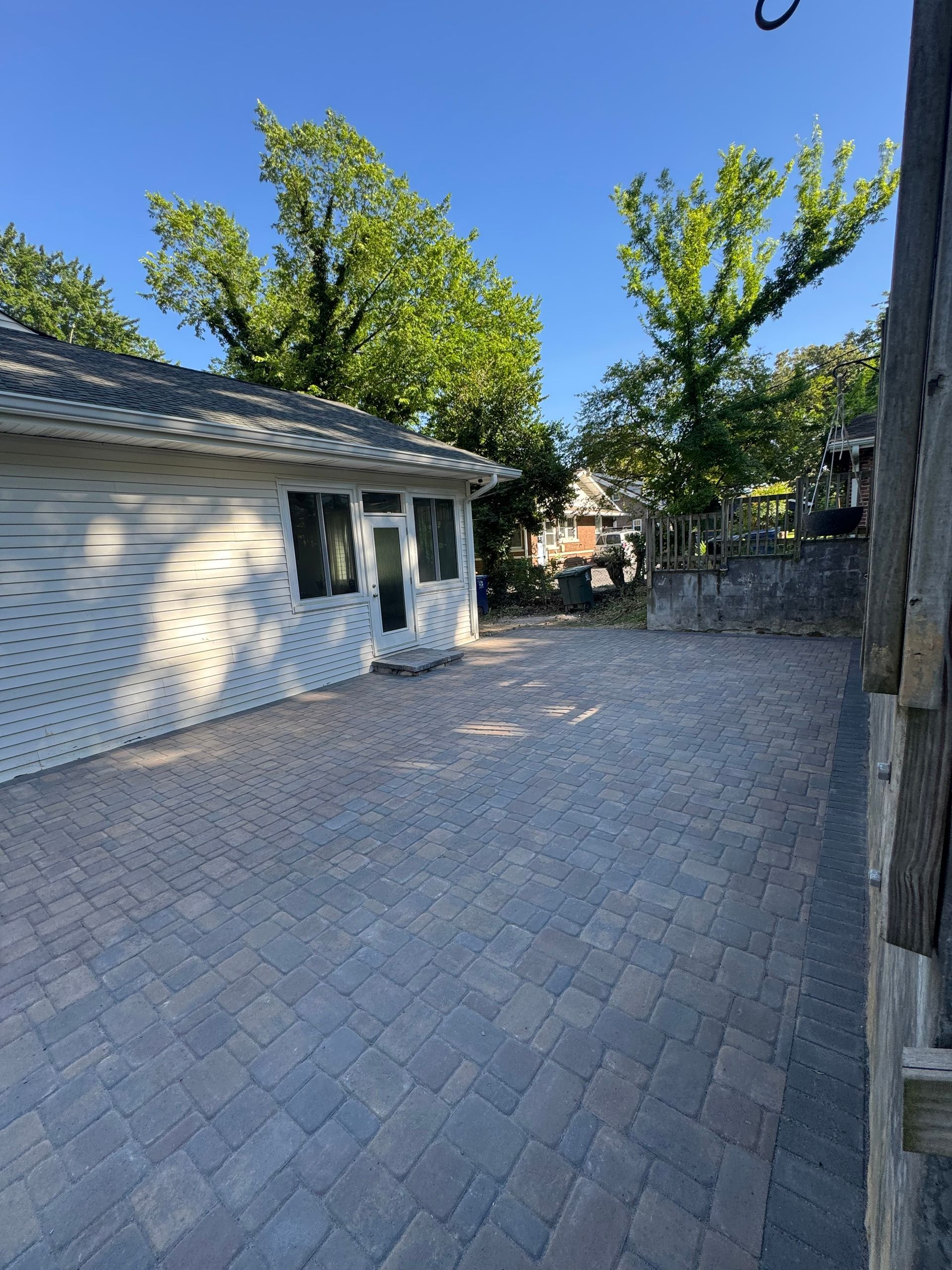 Paved backyard patio beside a white garage, with trees and a wooden fence in bright sunlight