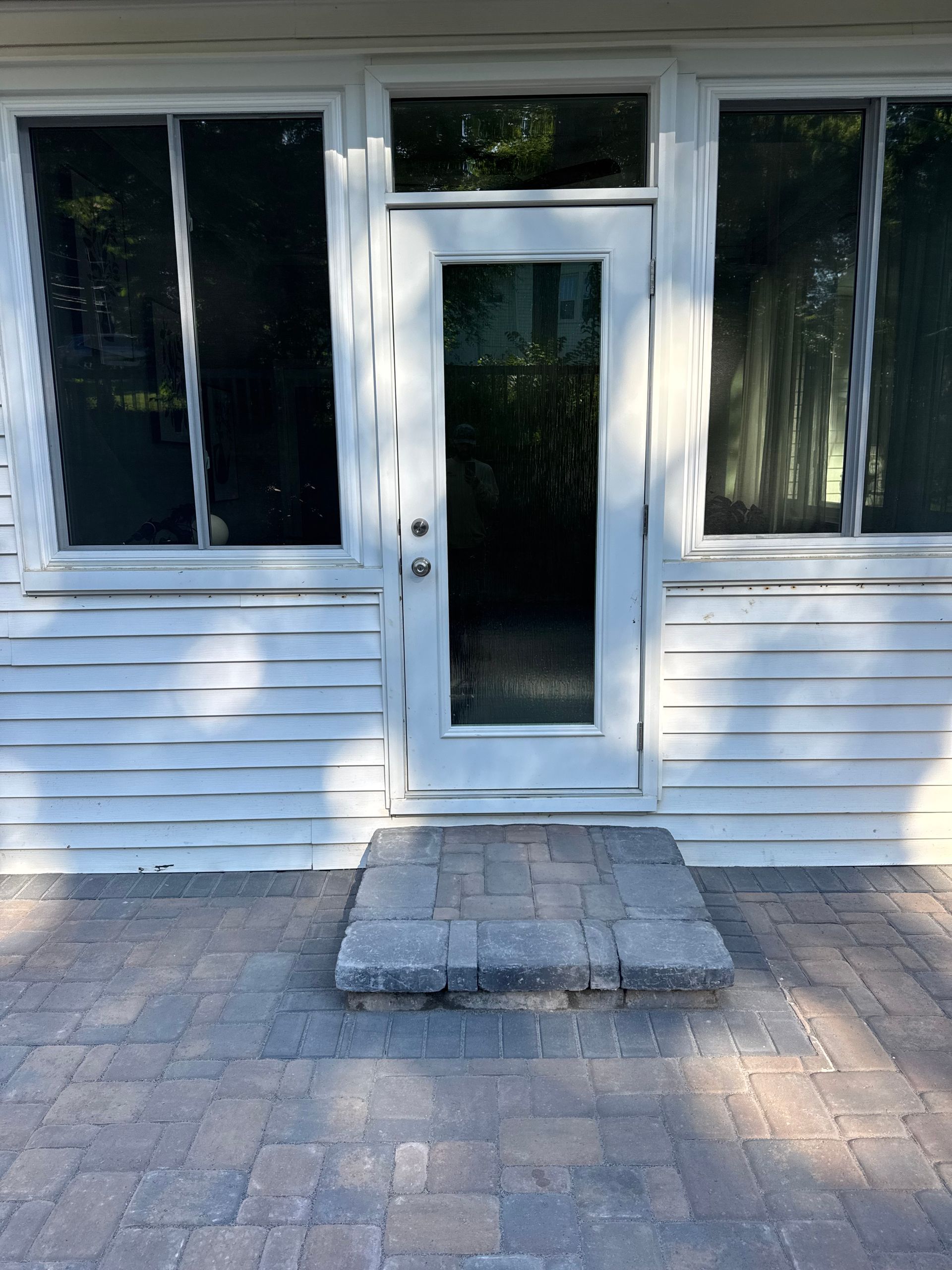 White house entrance with a glass door, two windows, and a small concrete step on a brick patio.
