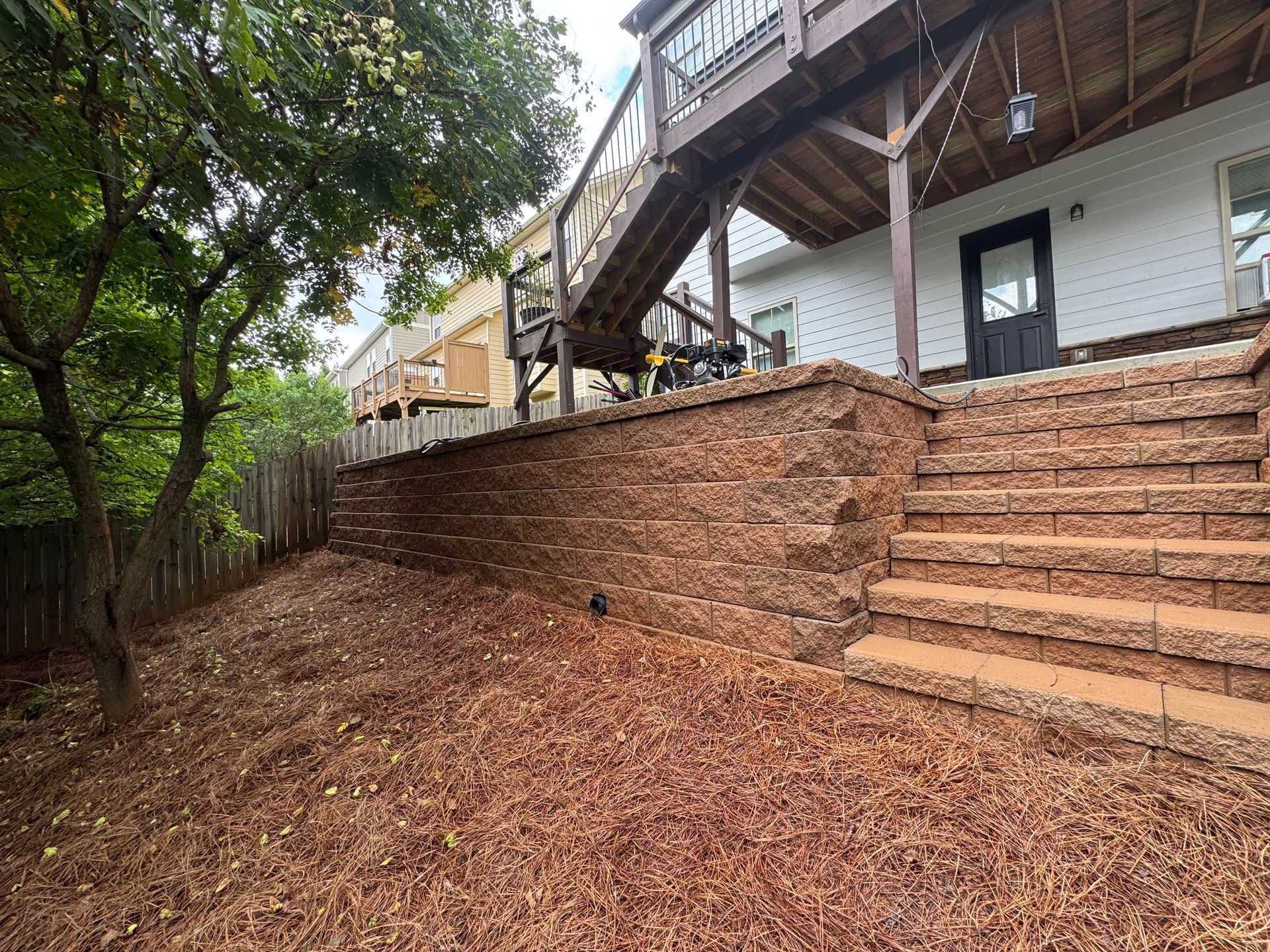 Steep wooded backyard with brick retaining wall, stairs, and an elevated deck beside a house