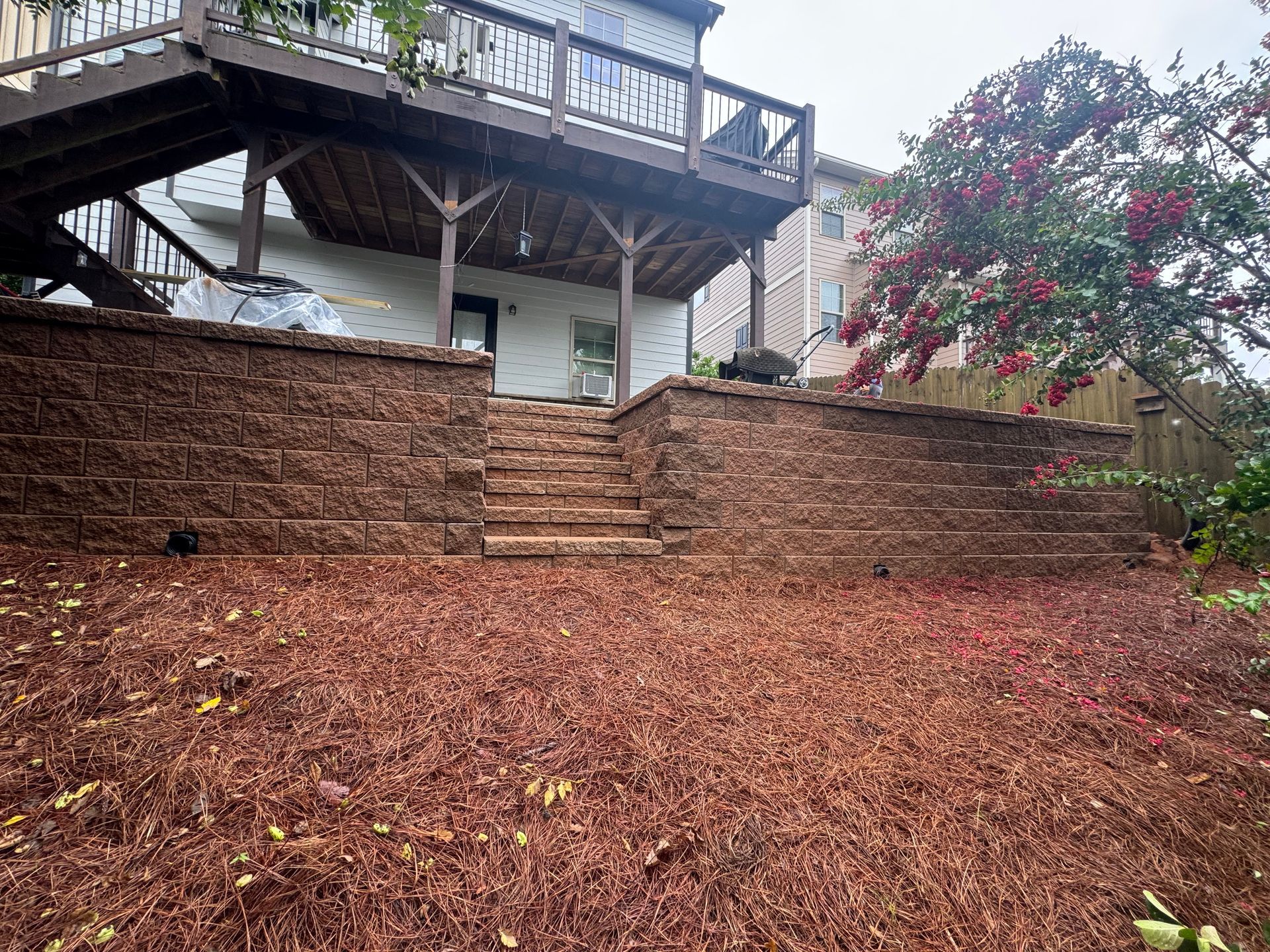 Backyard deck above a terraced retaining wall and leaf-covered ground, with a flowering tree on the right