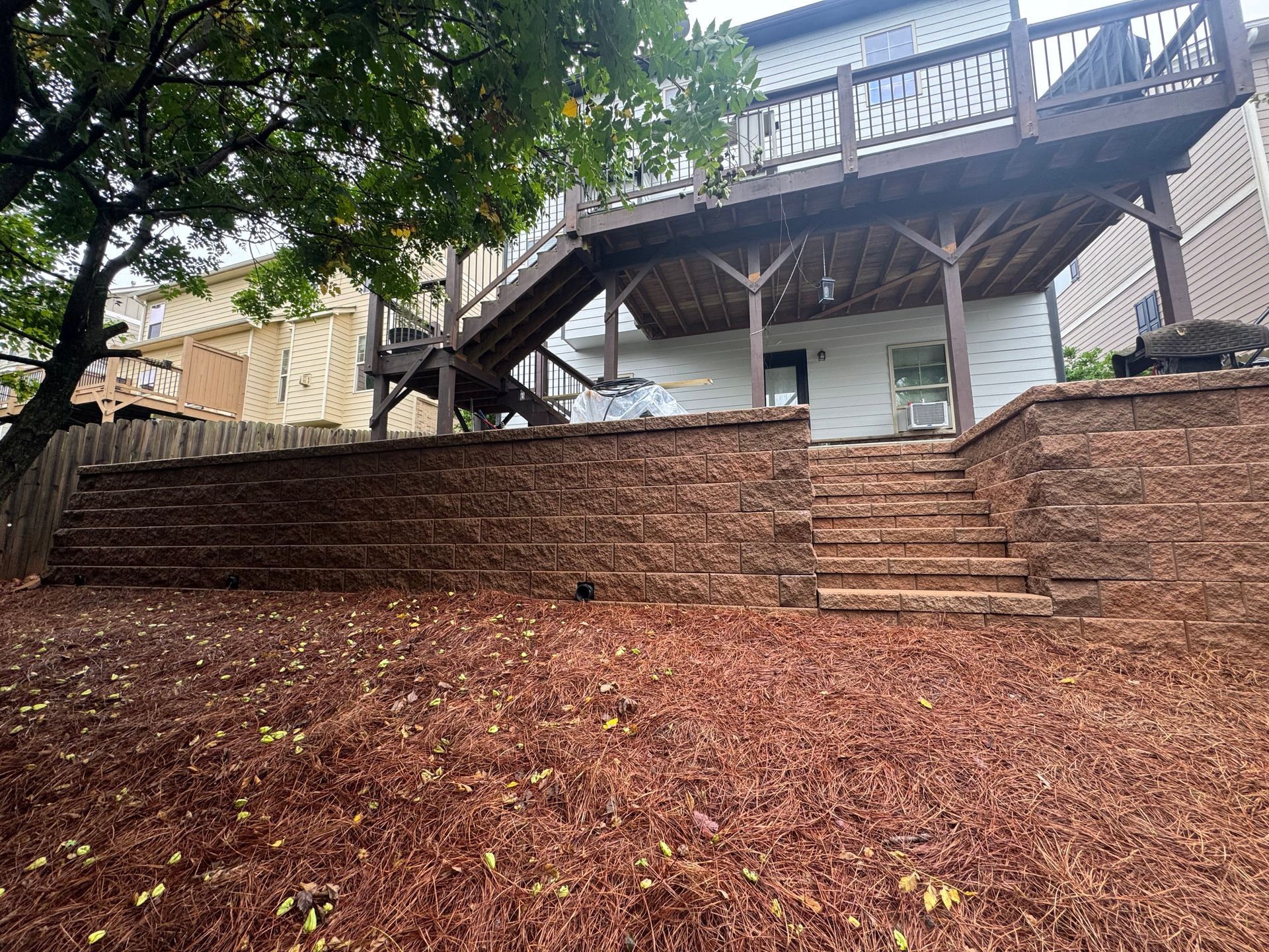 Brick retaining wall and steps leading to a wooden deck beside a house, with trees and a leaf-covered yard.