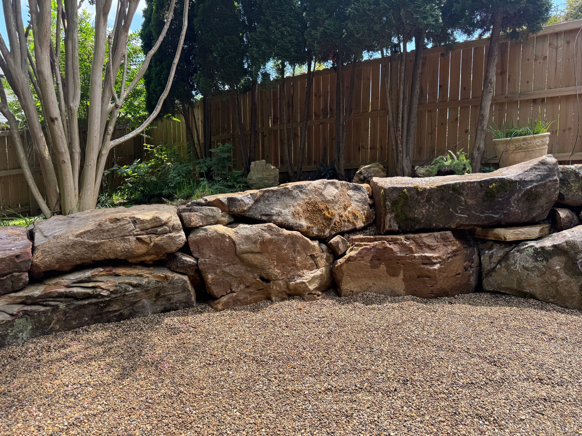 Stone retaining wall beside a gravel yard, with trees and a wooden fence in the background