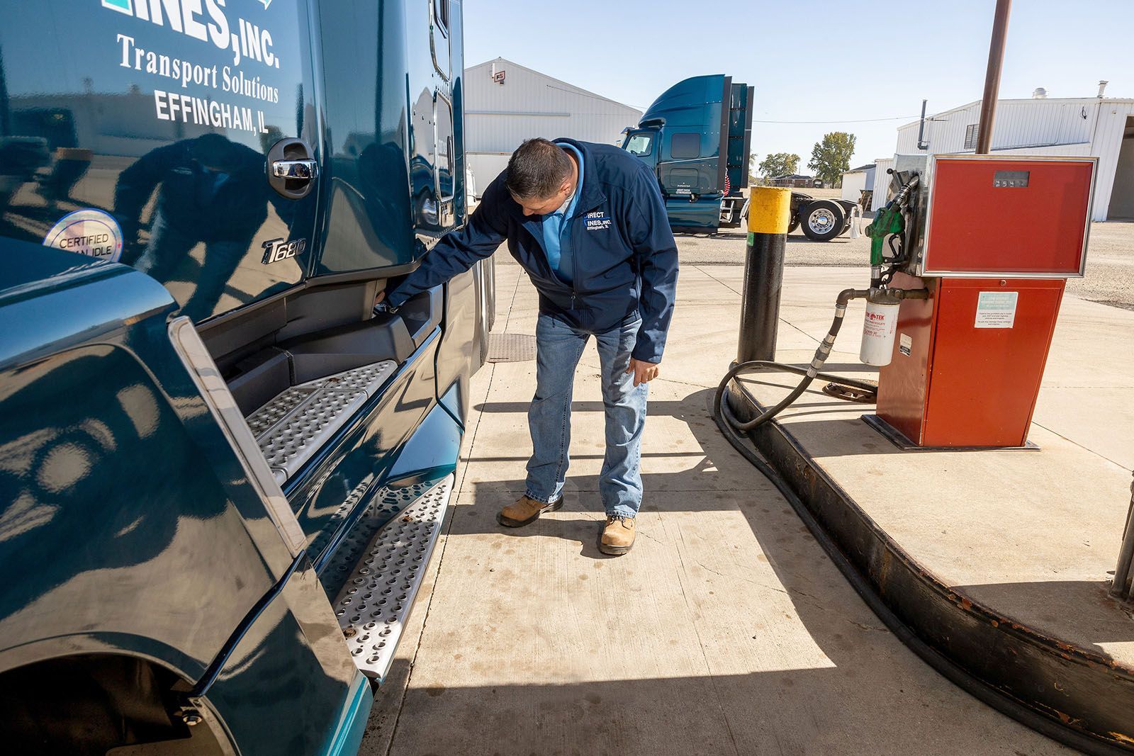 A person in blue jacket and jeans checks a truck at a gas pump