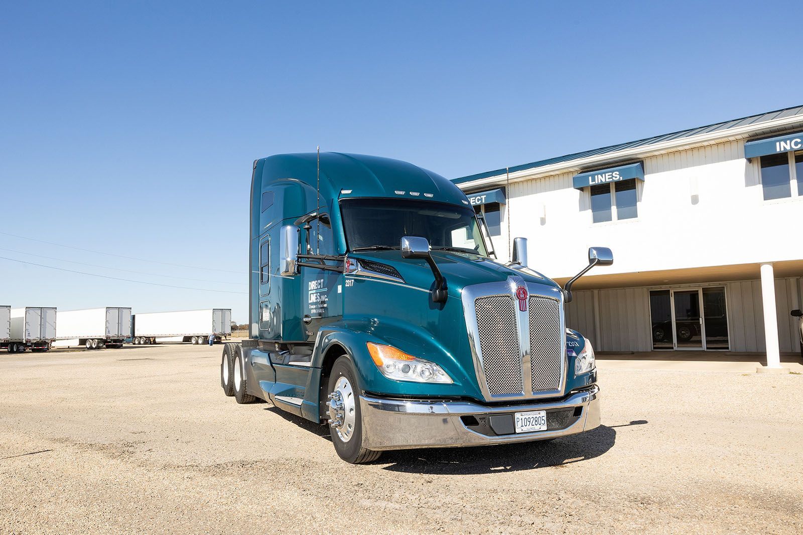 Teal semi-truck parked on gravel lot beside a white building