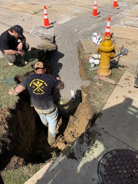 Men working on a trench near a fire hydrant; one is in the trench. Orange cones mark the area.