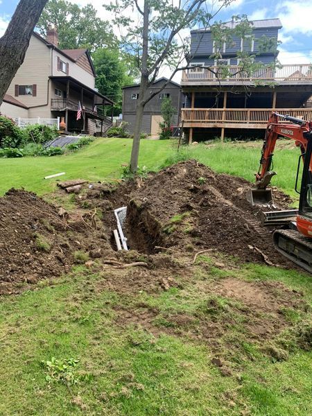 A back yard with an excavator digging a trench, dirt pile, and two houses in background.