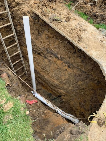 Dug trench with ladder and vertical white pipe. Gray pipe runs along bottom. Earth walls surround.