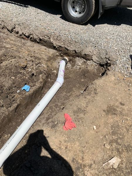 White PVC pipe in a dirt trench, near gravel and a vehicle tire.