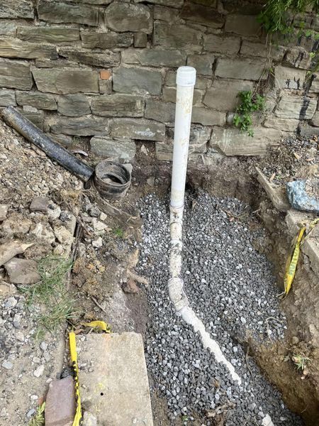 Trench with white and black pipes surrounded by gravel, next to a stone wall.