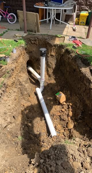 Trench dug with white PVC pipes installed. Dirt and tools surround the pipes in a yard setting.