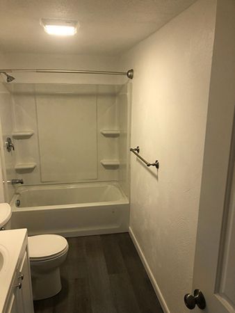 Bathroom with white walls, bathtub with glass door, toilet, and dark wood-look flooring.