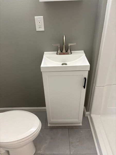 Small white bathroom vanity with sink and faucet; toilet on the left, grey wall, and grey floor.