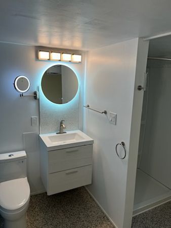 White bathroom with a floating vanity, round mirror, and a small shower.