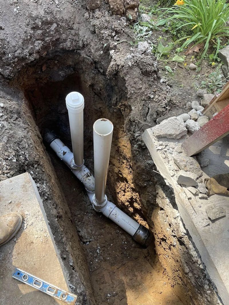 Pipes in a trench. White PVC pipes connected to a gray Y-shaped fitting. Dirt and concrete surround the pipes.