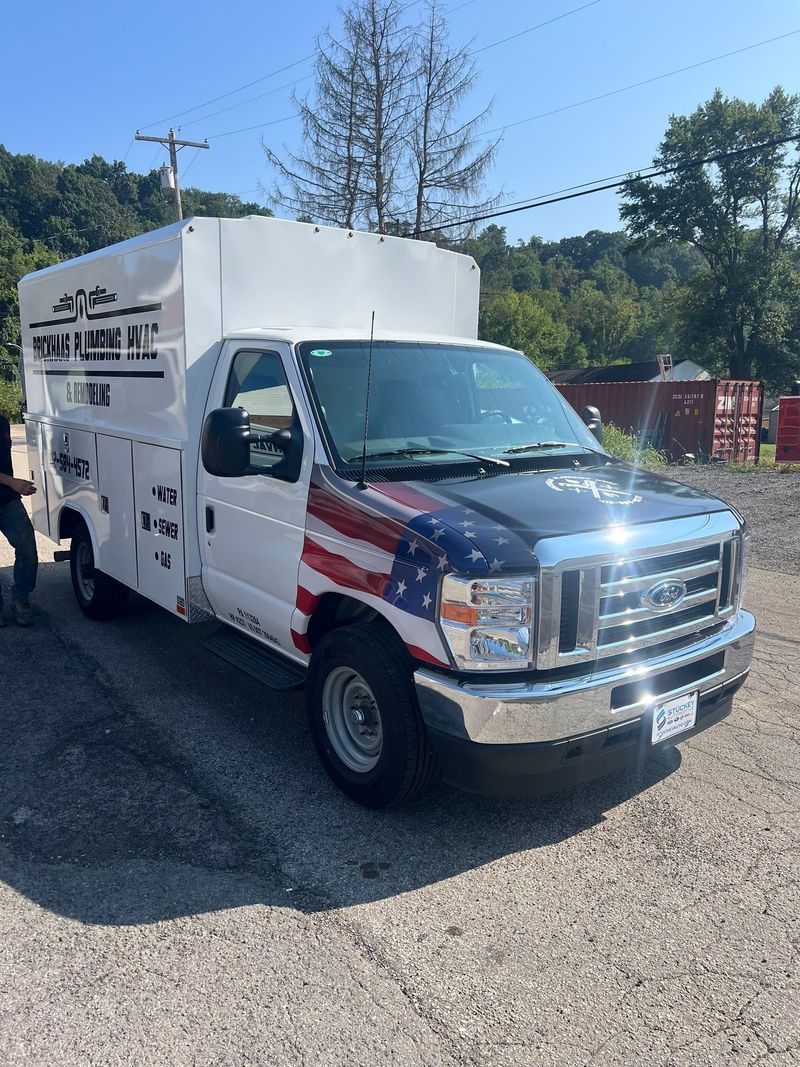 White utility truck with American flag design on the hood. Parked on gravel, trees in the background.