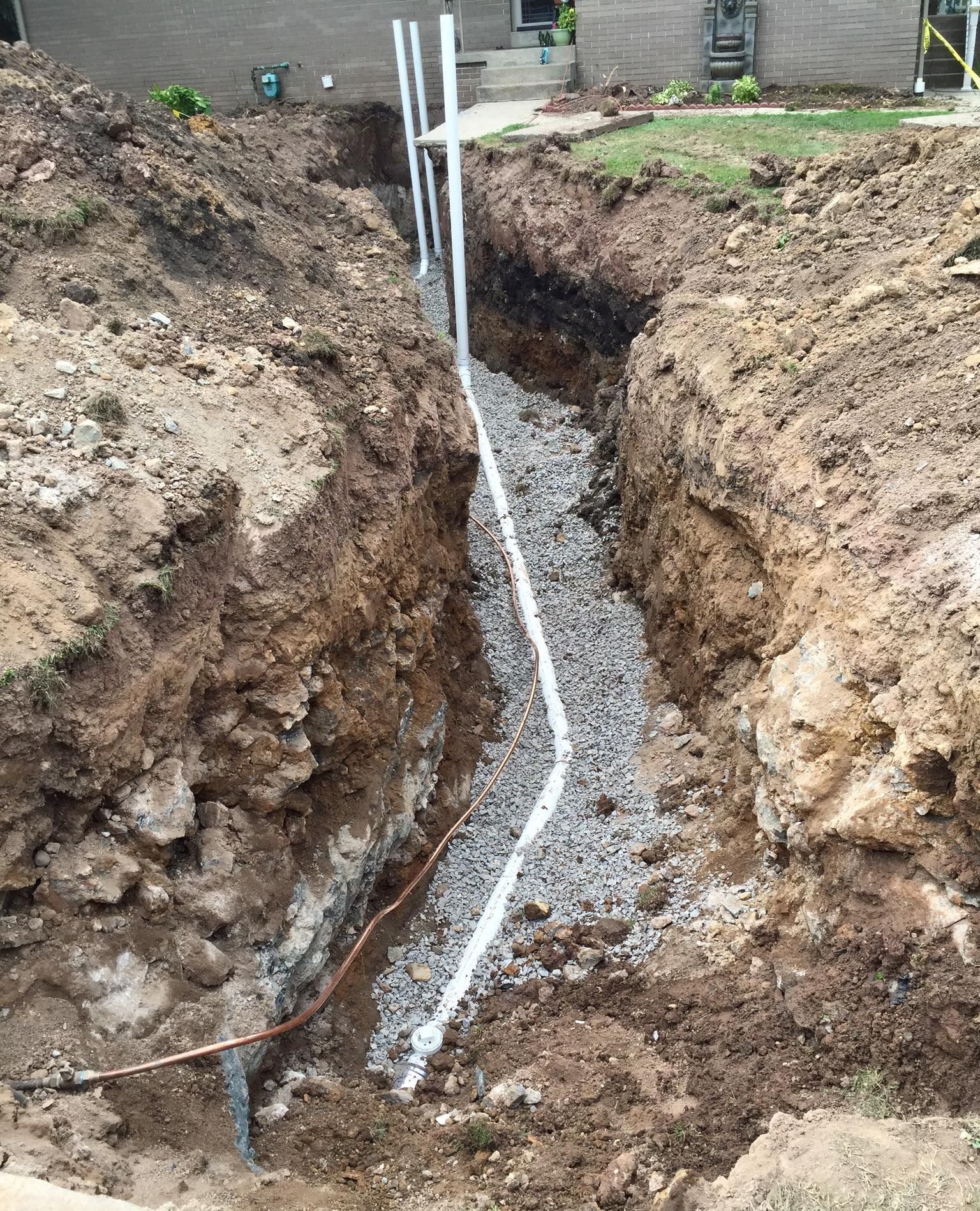 Trench with white pipe and gravel. Soil and grass in background.