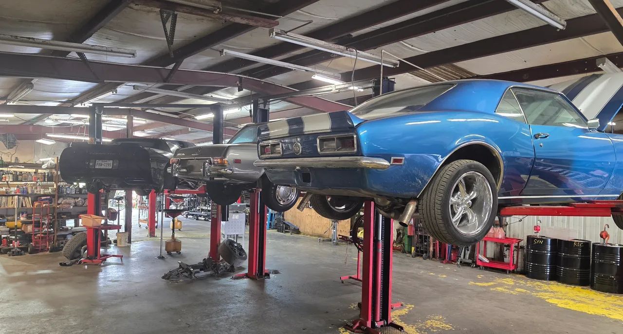 Blue car lifted on red hydraulic hoists in an auto repair shop garage with tools and equipment nearby