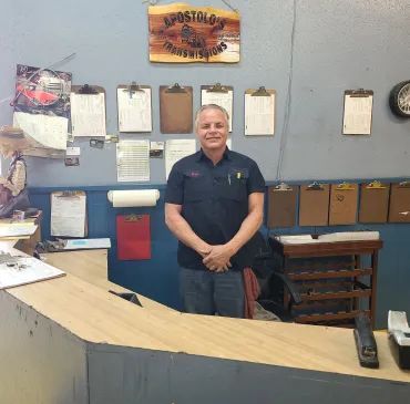 Man standing behind a counter in a small office with clipboards and shelves on the wall