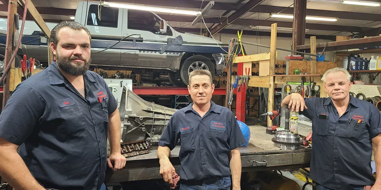 Three mechanics in blue uniforms stand in an auto repair shop beside a raised car.