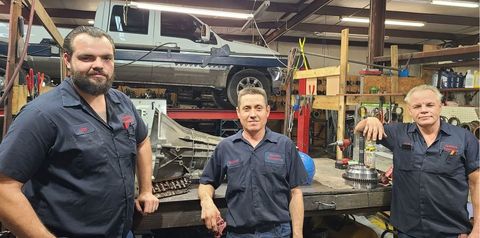Three mechanics in dark shirts standing in an auto repair shop with a car lifted on a hoist.