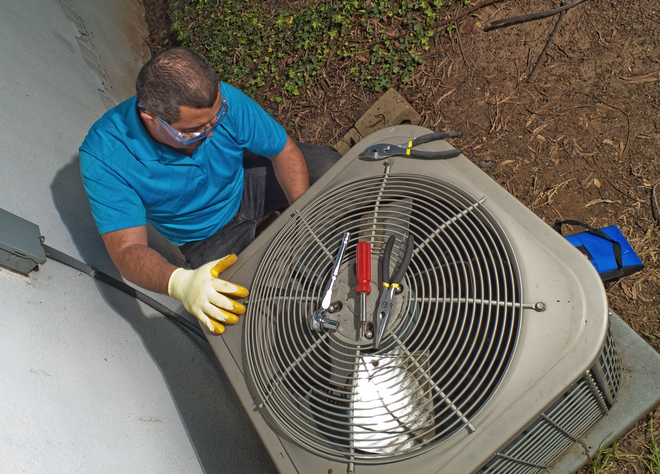 HVAC technician in blue shirt and safety glasses works on an outdoor air conditioning unit.