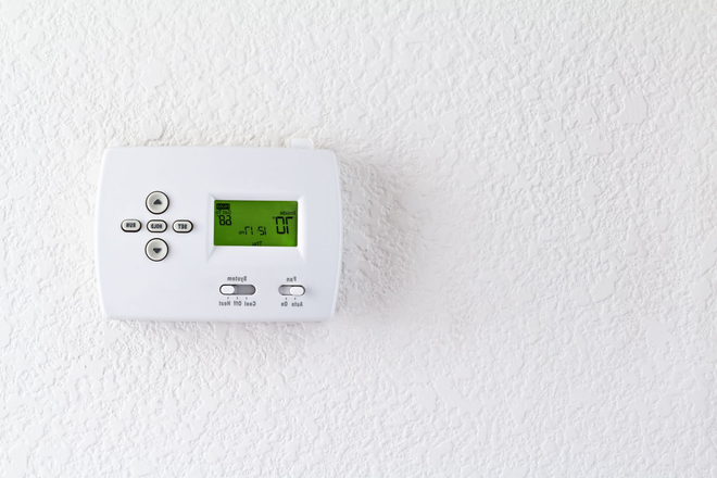 White thermostat mounted on a textured white wall; green display with temperature and music note icon visible.