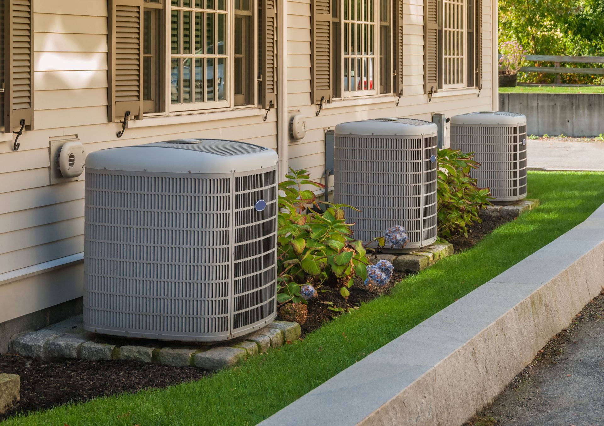 Three outdoor air conditioning units next to a white house with brown shutters and trim, set in a grassy area.