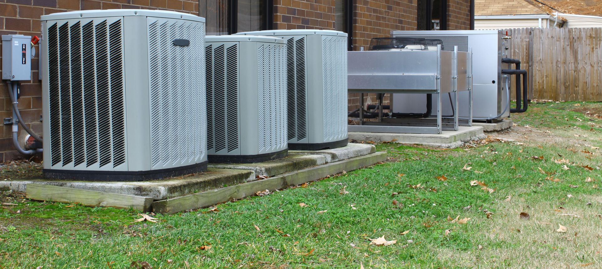 Air conditioning units on a concrete pad next to a brick building and a wooden fence.