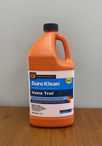 Orange SureKlean Vana Trol cleaner bottle on a wooden table against a gray wall