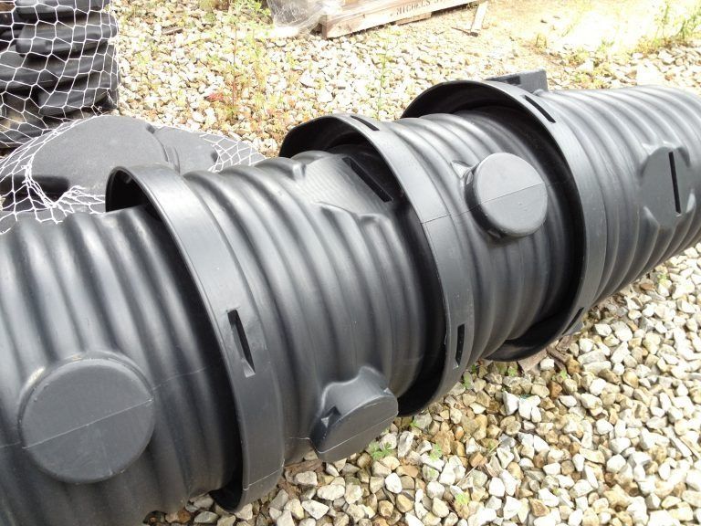 Black corrugated plastic drainage pipe with circular ports, lying on gravel outdoors
