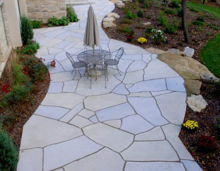 Stone patio with a metal table and umbrella, bordered by landscaping and a winding walkway
