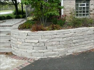 Curved white stone retaining wall with plants beside a driveway and sidewalk near a house